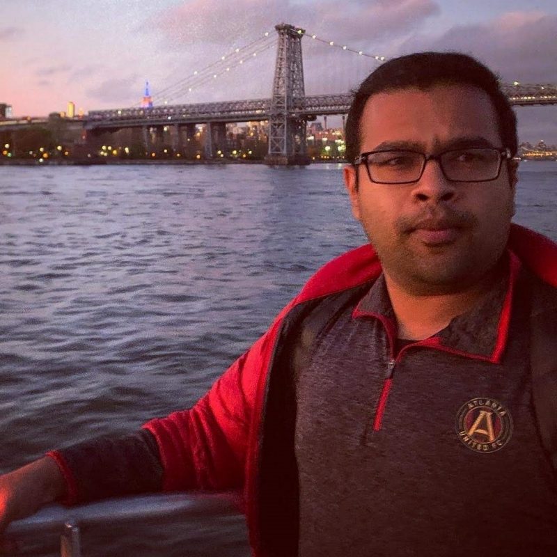 Maruf poses confidently for a picture in featuring a lovely bridge crossing a river in a sunset scene, wearing an unzipped jacket