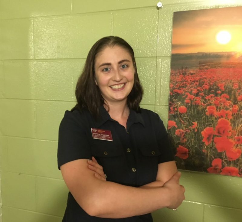Martina stands smiling at the camera, in front of a green-painted wall with a large photo of a field of flowers. She is in the gender-neutral bathroom on the first floor of the GLC