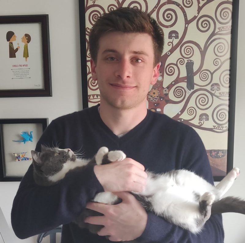 Jack smiles calmly into the camera, standing before an image of a family tree styled after yggdrasil. His befuddled cat is cradled in his arms. Jack's hair is brown. The cat is white and multi-tones of grey.