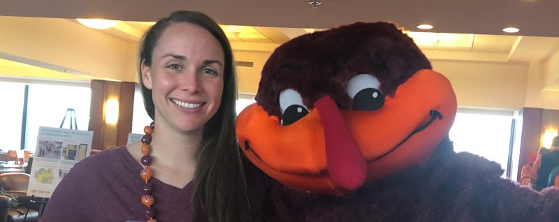 a female graduate student faces the camera, smiling, arm in arm with the Hokie Bird